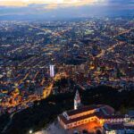 La imagen muestra una vista nocturna de una gran ciudad llena de luces que se extienden hasta el horizonte. En primer plano, sobre una montaña, hay una iglesia iluminada con techo rojizo y una torre blanca. El cielo tiene tonos azules y naranjas del atardecer.