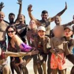 Grupo de influencers en la playa, cubiertas de lodo, posando sonrientes frente al mar.